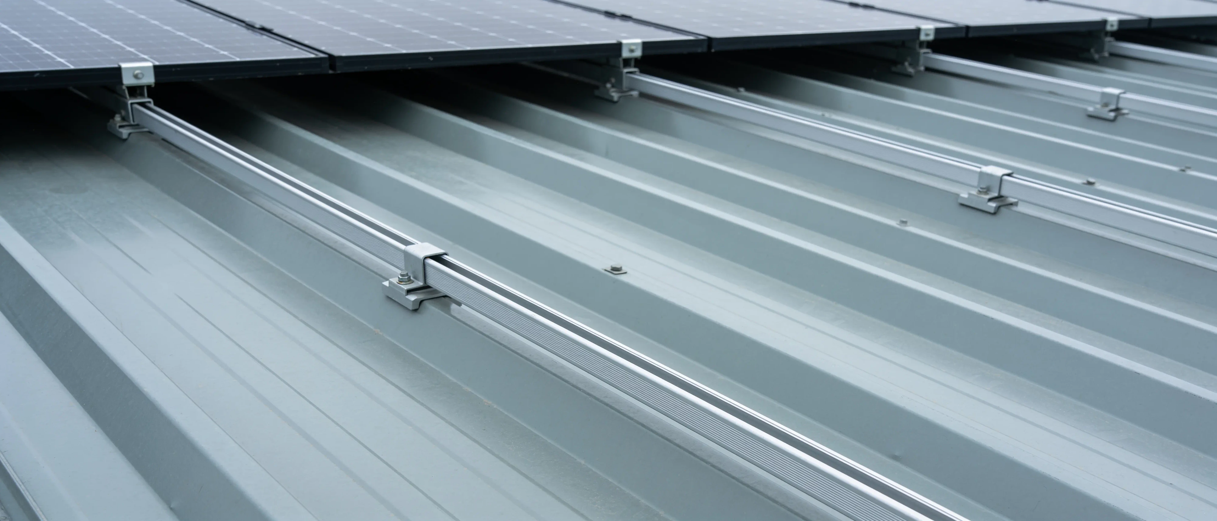 Montageschienen aus Aluminium auf einem hellgrauen Trapezblechdach, darüber der Rand eines Solarmoduls